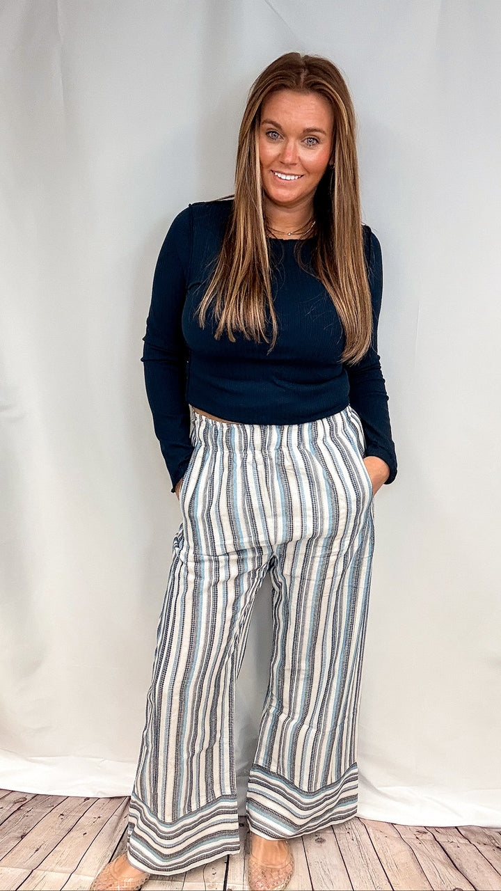 A woman modeling a dark navy long-sleeve top and high-waisted, wide-leg striped pants with vertical stripes in navy, gray, and white, standing against a white studio backdrop.