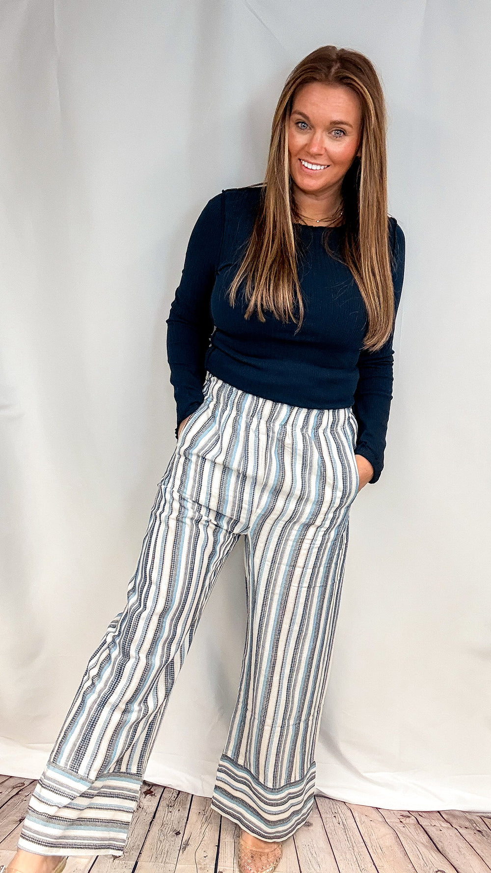 A woman modeling a pair of high-waisted, wide-leg pants with vertical blue, gray, and white stripes, paired with a dark navy long-sleeve top, standing against a white fabric backdrop.