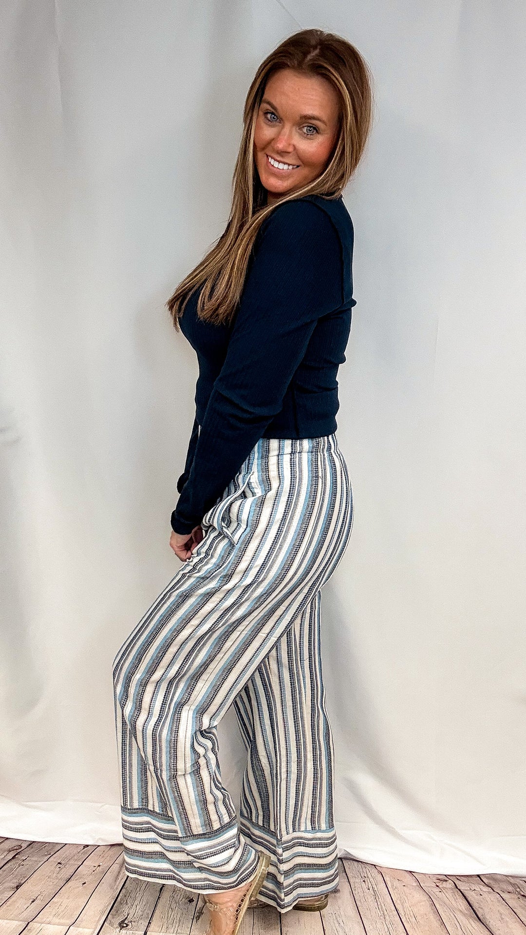 A woman modeling a navy blue long-sleeve top and high-waisted, wide-leg striped pants with vertical stripes in navy, beige, and white, standing against a white studio backdrop.