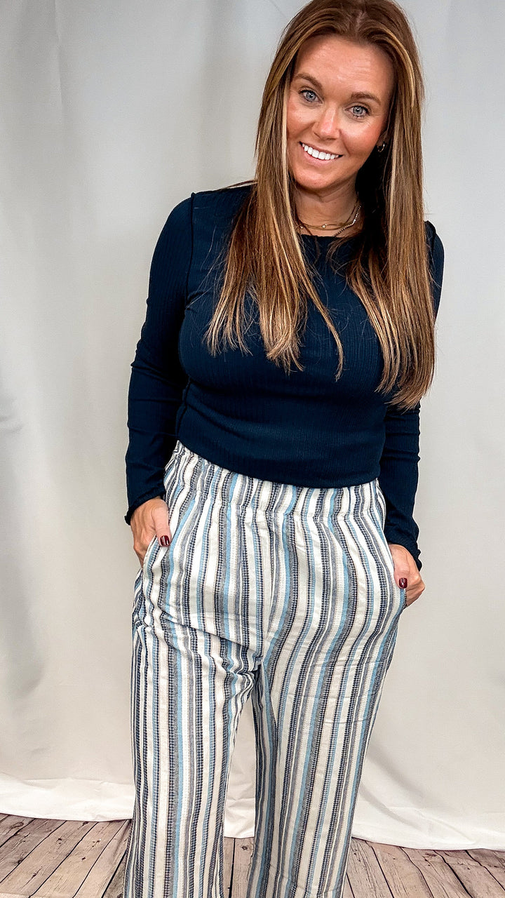 A woman modeling a dark navy long-sleeve top and high-waisted, wide-leg trousers with vertical blue, gray, and white stripes, standing against a white studio backdrop.