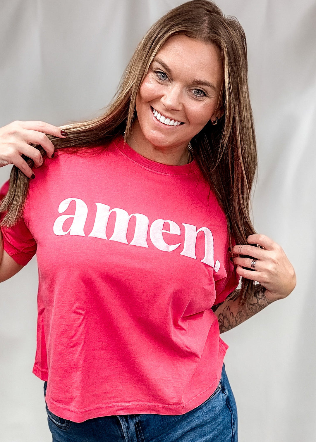 A smiling woman with long brown hair wears a bright pink short-sleeve t-shirt with the word &