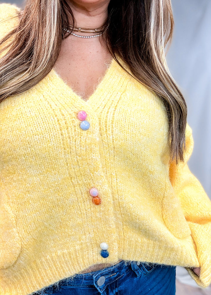 A close-up of a woman wearing a fuzzy yellow knit cardigan with colorful pom-pom buttons, paired with blue jeans.