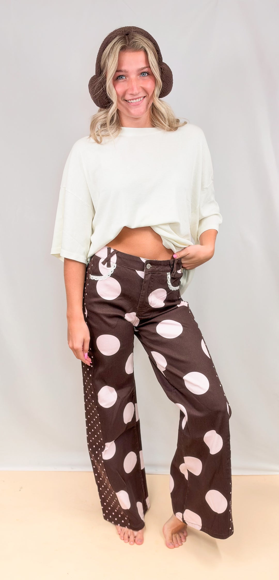 Woman wearing brown pants with large pink polka dots on them and cream top on plain background