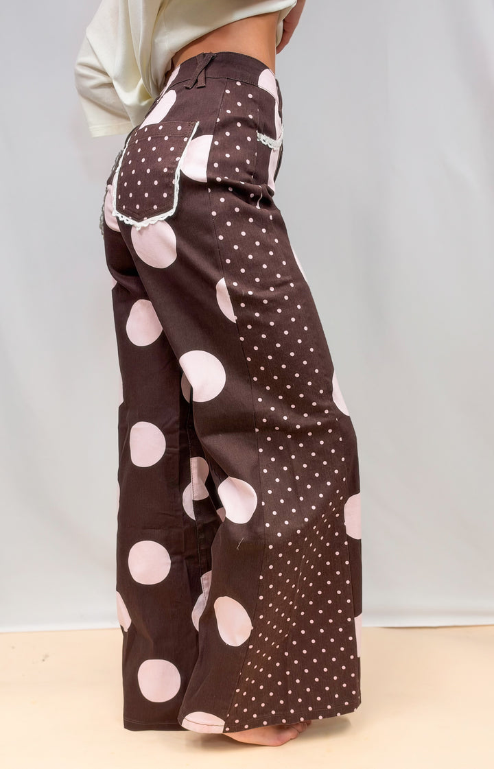 Woman shown from side view wearing brown pants with large and small light pink polka dots on plain background
