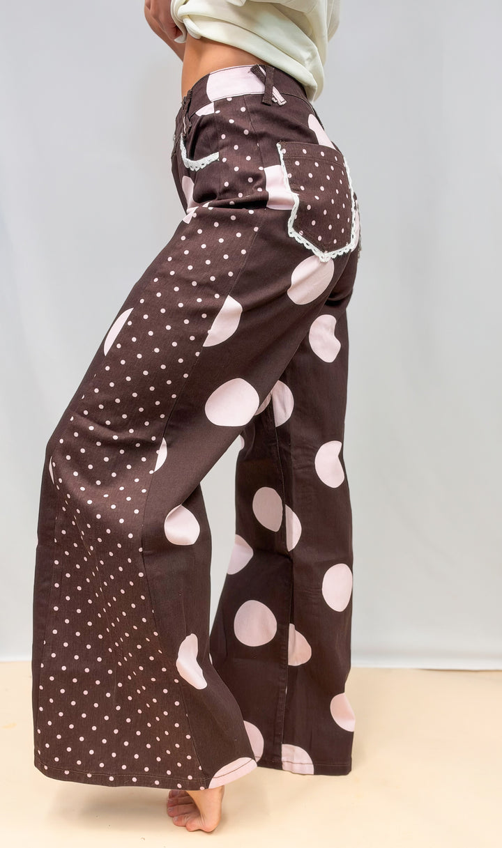 Woman shown from side view wearing brown pants with large and small light pink polka dots on plain background