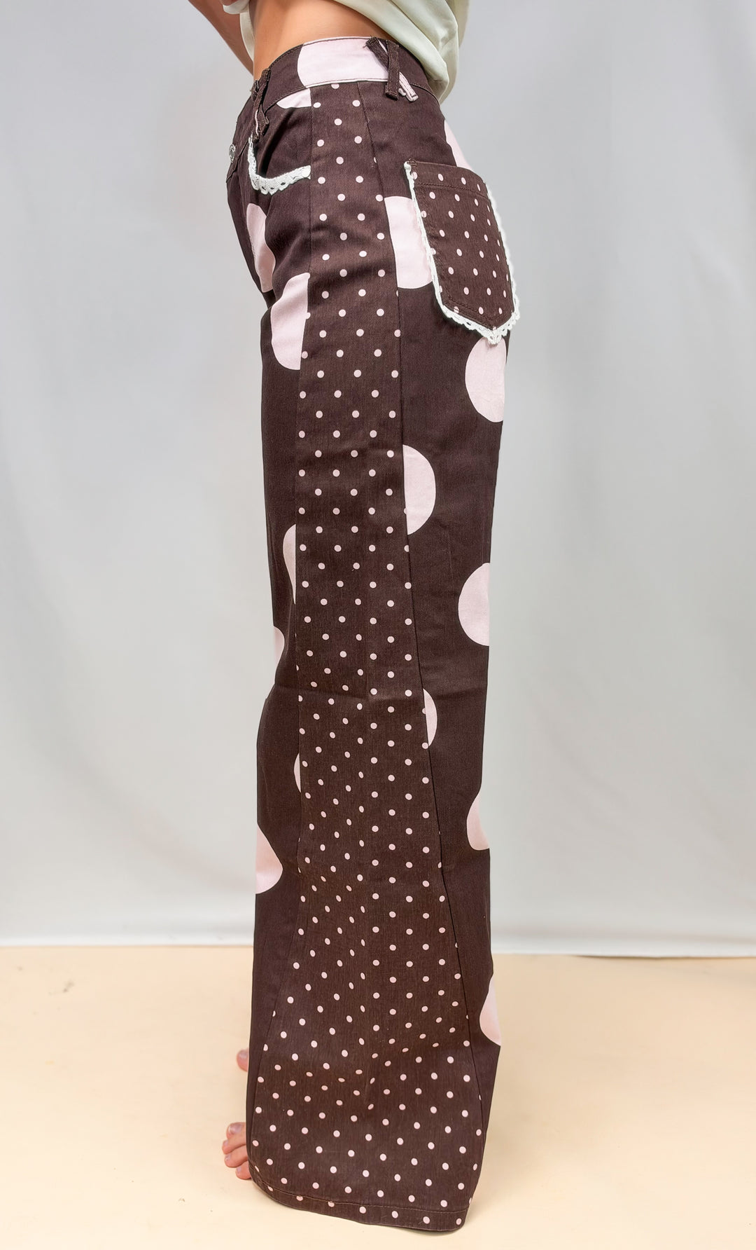 Woman shown from side view wearing brown pants with large and small light pink polka dots on plain background