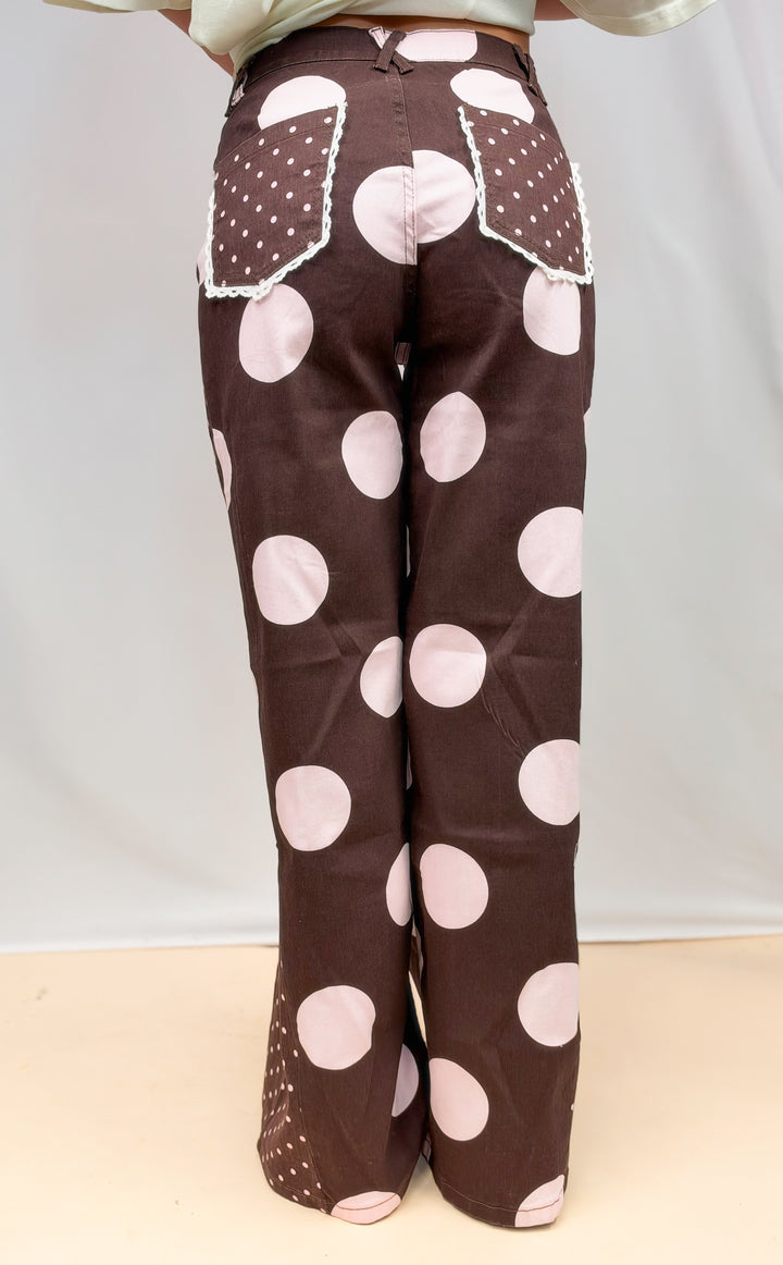 Woman shown from back wearing brown pants with large light pink polka dots on plain background