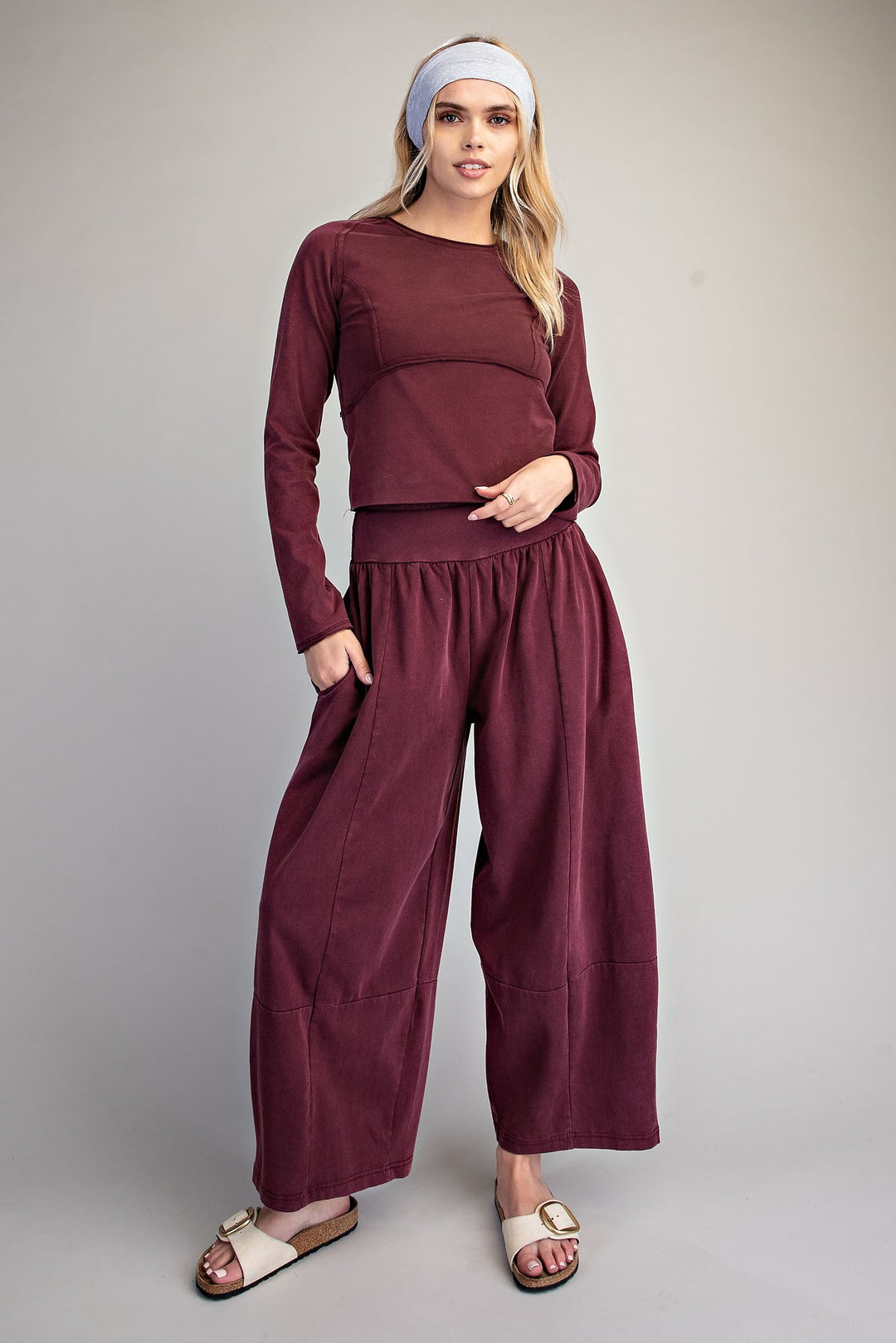 A woman modeling a matching burgundy two-piece lounge set consisting of a long-sleeve cropped top and wide-leg pants, styled with a white headband and beige slide sandals against a neutral gray studio background.