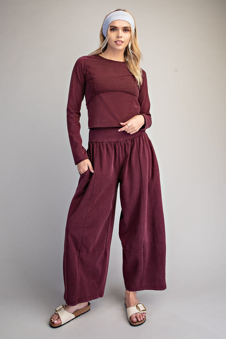 A woman modeling a matching burgundy two-piece lounge set consisting of a long-sleeve cropped top and wide-leg pants, styled with a white headband and beige slide sandals against a neutral gray studio background.