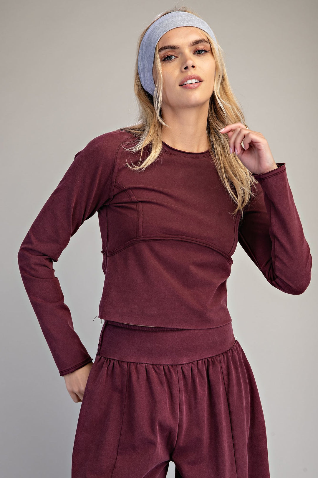 A woman modeling a matching burgundy long-sleeve crop top and high-waisted pants set, paired with a light blue-gray headband, against a neutral gray studio background.