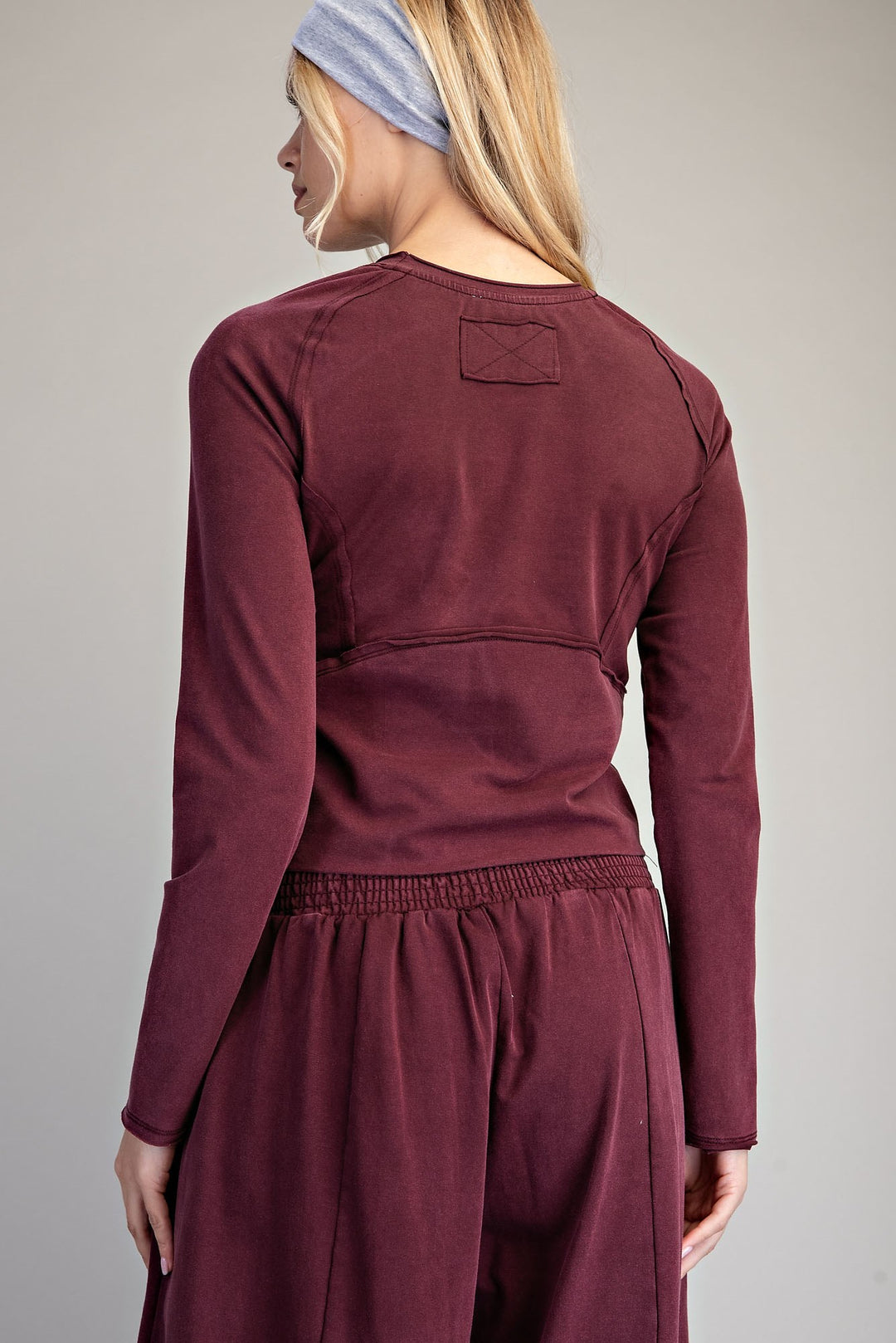 A woman shown from the back wearing a long-sleeve maroon scrub top with a shoulder patch and matching scrub pants against a neutral gray background.
