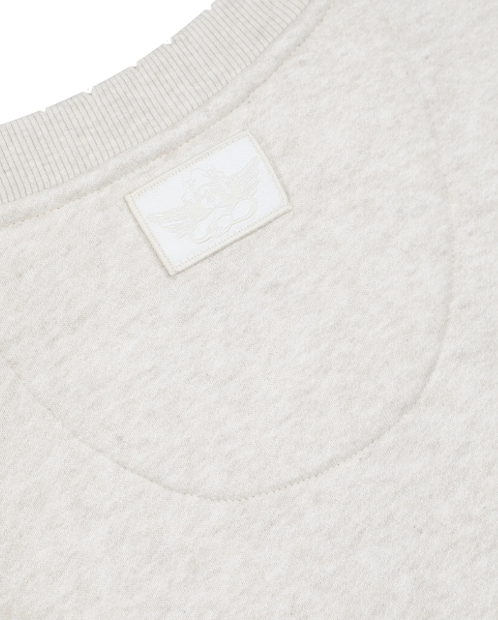 Close-up of a light cream knit garment showing the ribbed crew neck, a small rectangular brand patch, and the embroidered logo.