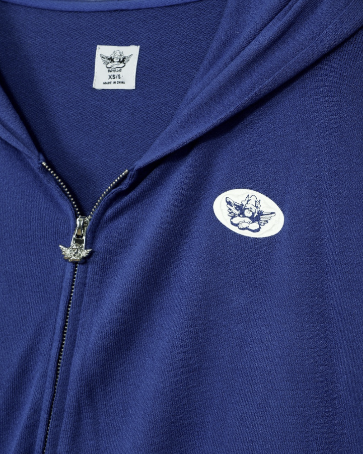 Close-up of a royal blue zip-up hoodie featuring a white chest patch with a black winged emblem, a white neck label, and a silver zipper pull.