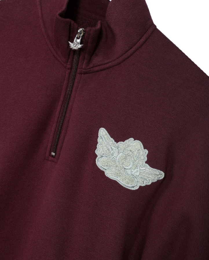 Close-up of a maroon quarter-zip pullover sweatshirt featuring a silver embroidered angel wing logo on the left chest and a silver zipper pull with a wing design.