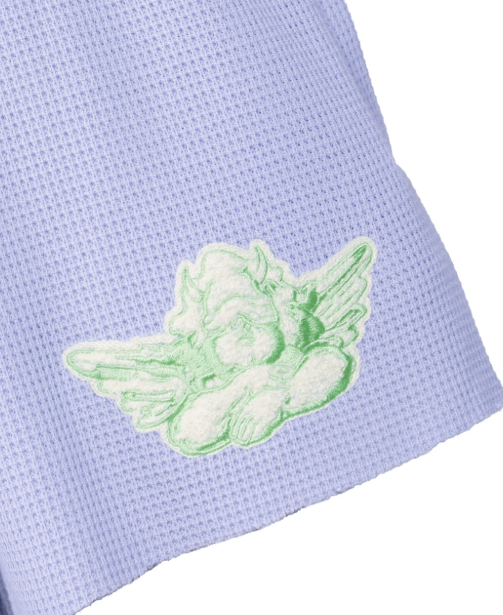 A close-up of a light lavender waffle-knit fabric with a light green embroidered cherub motif on the side.