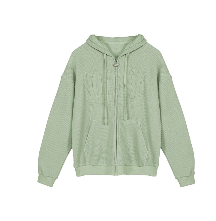A sage green zip-up hoodie with a textured waffle knit pattern, featuring a drawstring hood, front pockets, and a tonal embroidered &