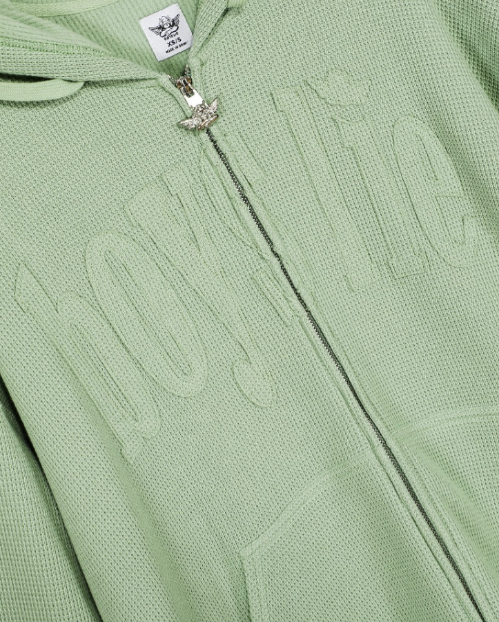 A close-up of a light sage green zip-up hoodie featuring a textured waffle knit fabric, a large tonal embroidered logo on the chest, a silver zipper, and a white brand label.
