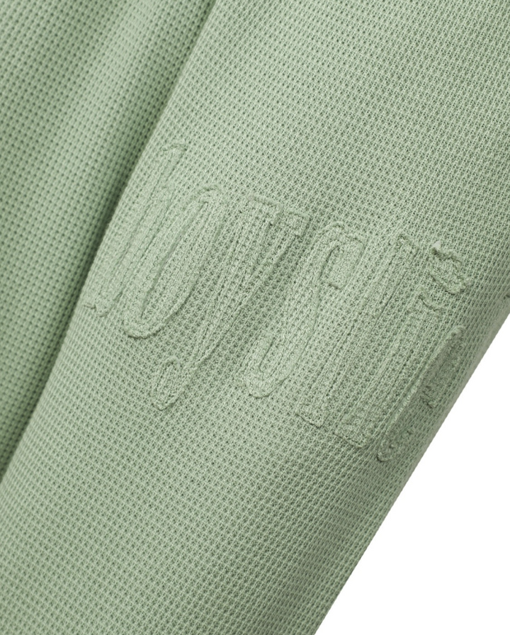 Close-up of a sage green textured fabric with an embossed tonal logo, showing the fine mesh-like weave and a sliver of white background at the bottom right.