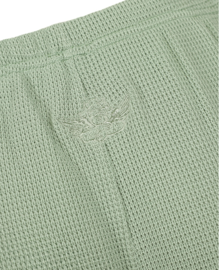 Close-up of a light sage green waffle-knit fabric with a tonal embroidered winged logo on the front.