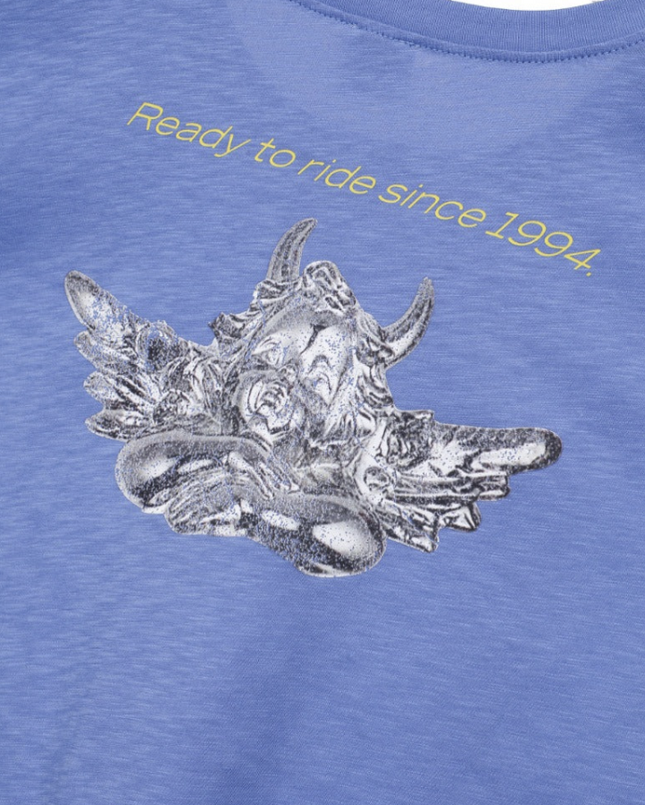 Close-up of a heathered periwinkle blue t-shirt featuring a metallic silver graphic of a horned motorcycle helmet with roses, and yellow text reading &