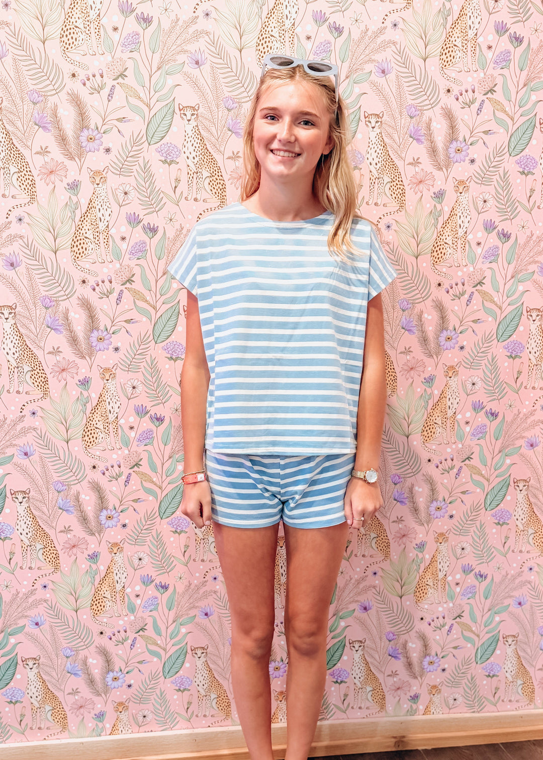 A woman stands barefoot in front of a pink patterned wallpaper wall, wearing a light blue and white striped t-shirt and matching striped shorts.