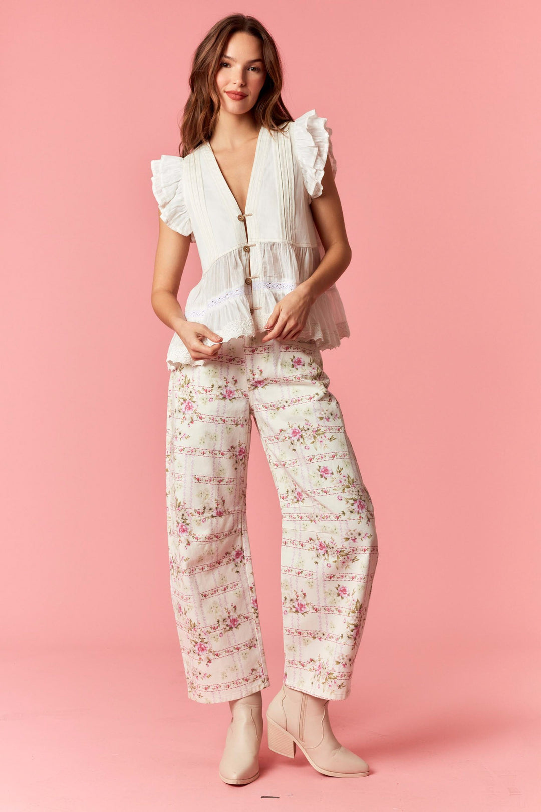 Woman wearing a white blouse with ruffled sleeves and floral pants on a pink background