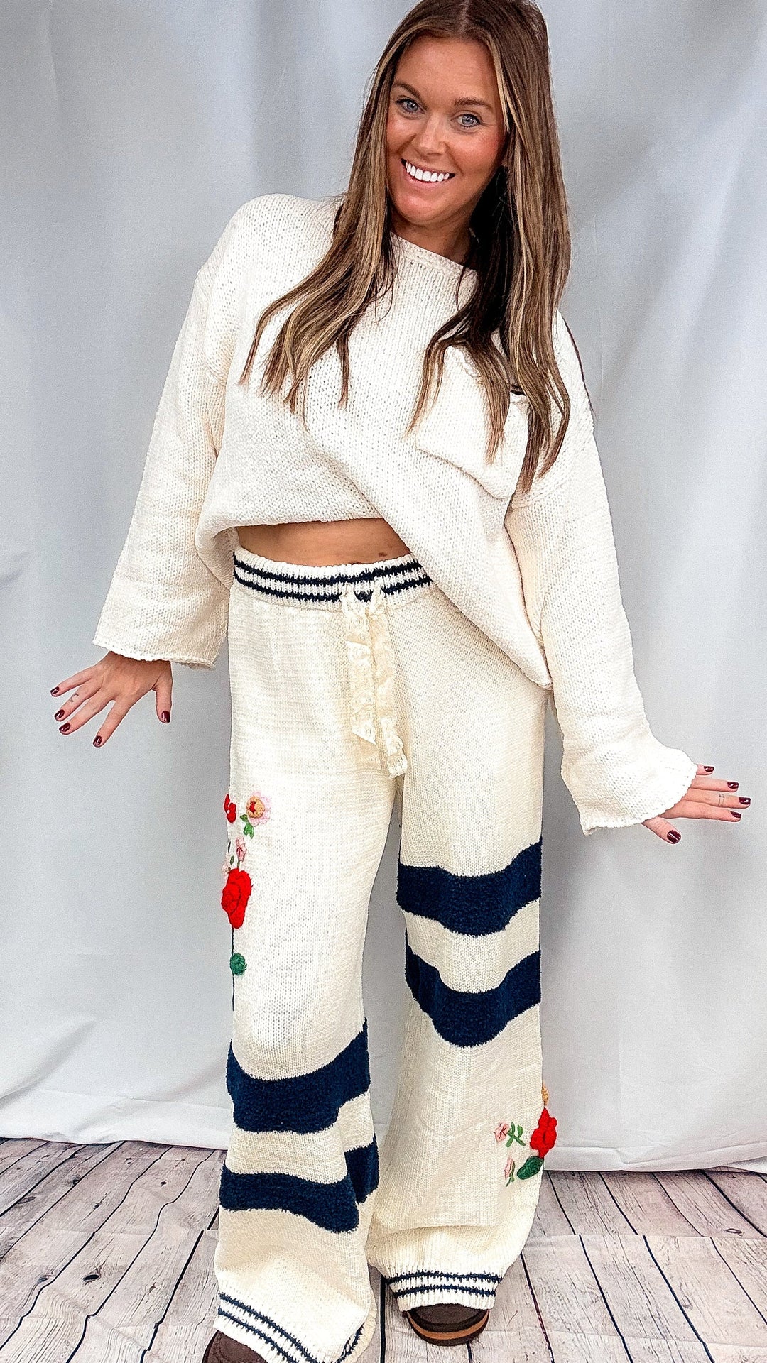 A woman modeling a cream-colored knit lounge set consisting of a cropped long-sleeve top and matching wide-leg pants with navy blue stripes and red floral embroidery, standing against a light gray backdrop.
