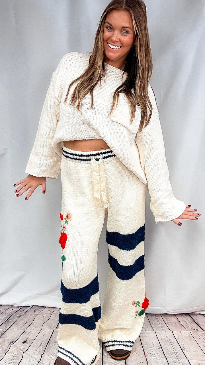 A woman modeling a cream-colored knit lounge set consisting of a cropped long-sleeve top and matching wide-leg pants with navy blue stripes and red floral embroidery, standing against a light gray backdrop.