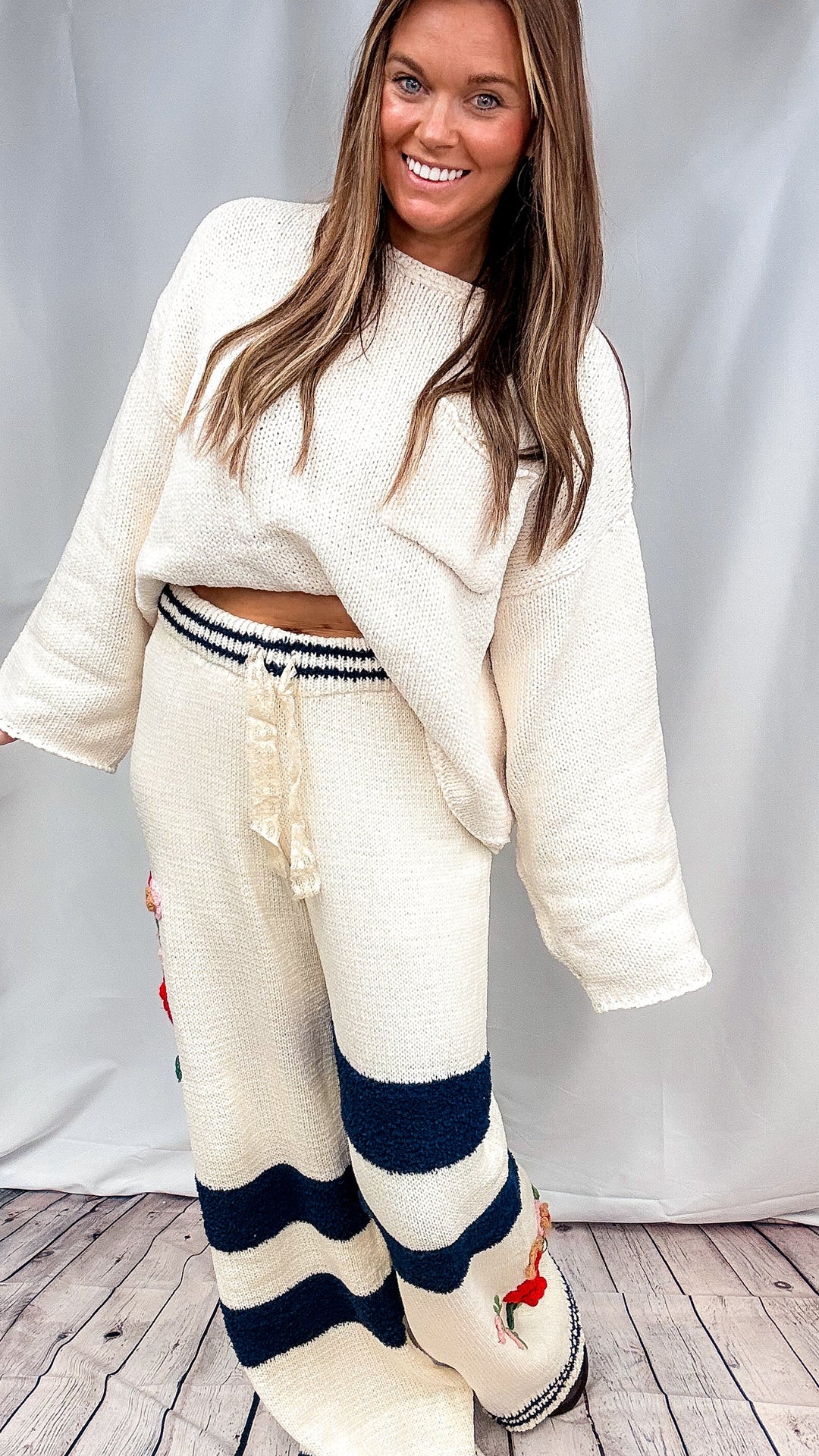 A woman modeling a cozy knit two-piece lounge set featuring a cream cropped sweater and matching wide-leg pants with navy and cream stripes, set against a white draped backdrop.