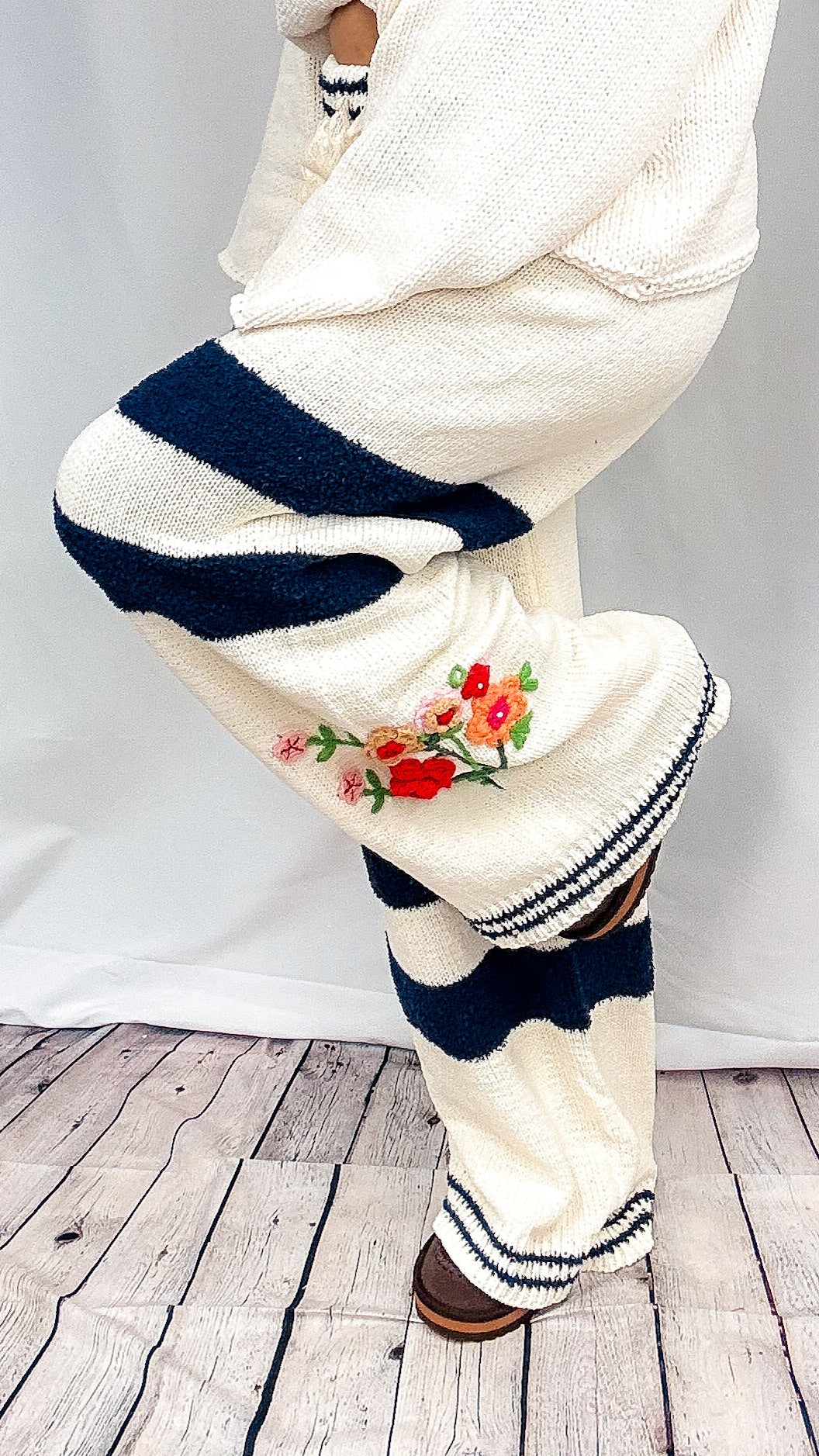 Cream knit pants with bold navy blue stripes, a red and green floral embroidery on the side, and striped ribbed cuffs, shown on a model against a white draped backdrop and wood floor.