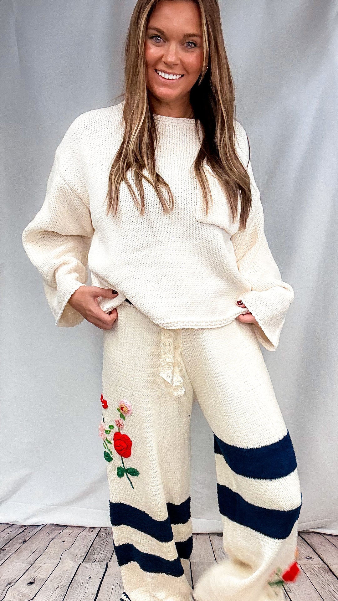 A woman modeling a cream-colored knit lounge set featuring a matching sweater and sweatpants. The sweatpants are decorated with navy blue horizontal stripes and a red rose graphic on the left leg, with striped cuffs.