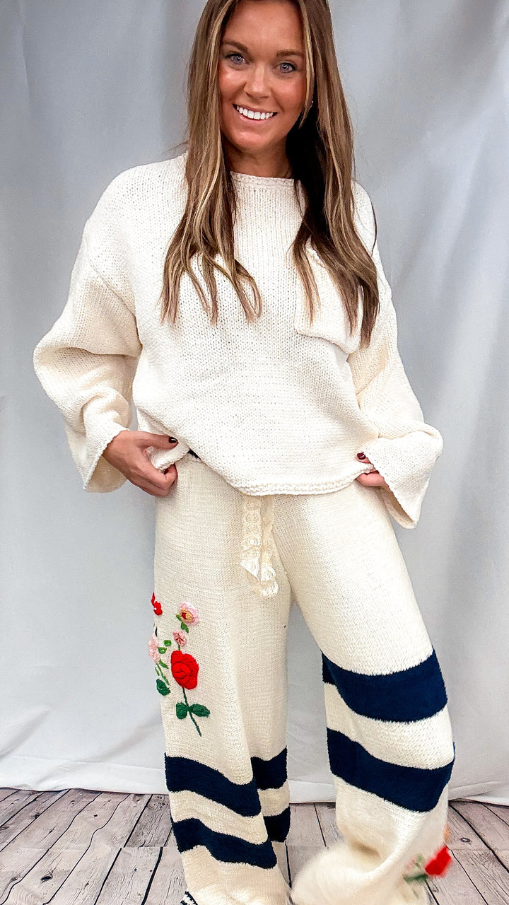 A woman modeling a cream-colored knit lounge set featuring a matching sweater and sweatpants. The sweatpants are decorated with navy blue horizontal stripes and a red rose graphic on the left leg, with striped cuffs.