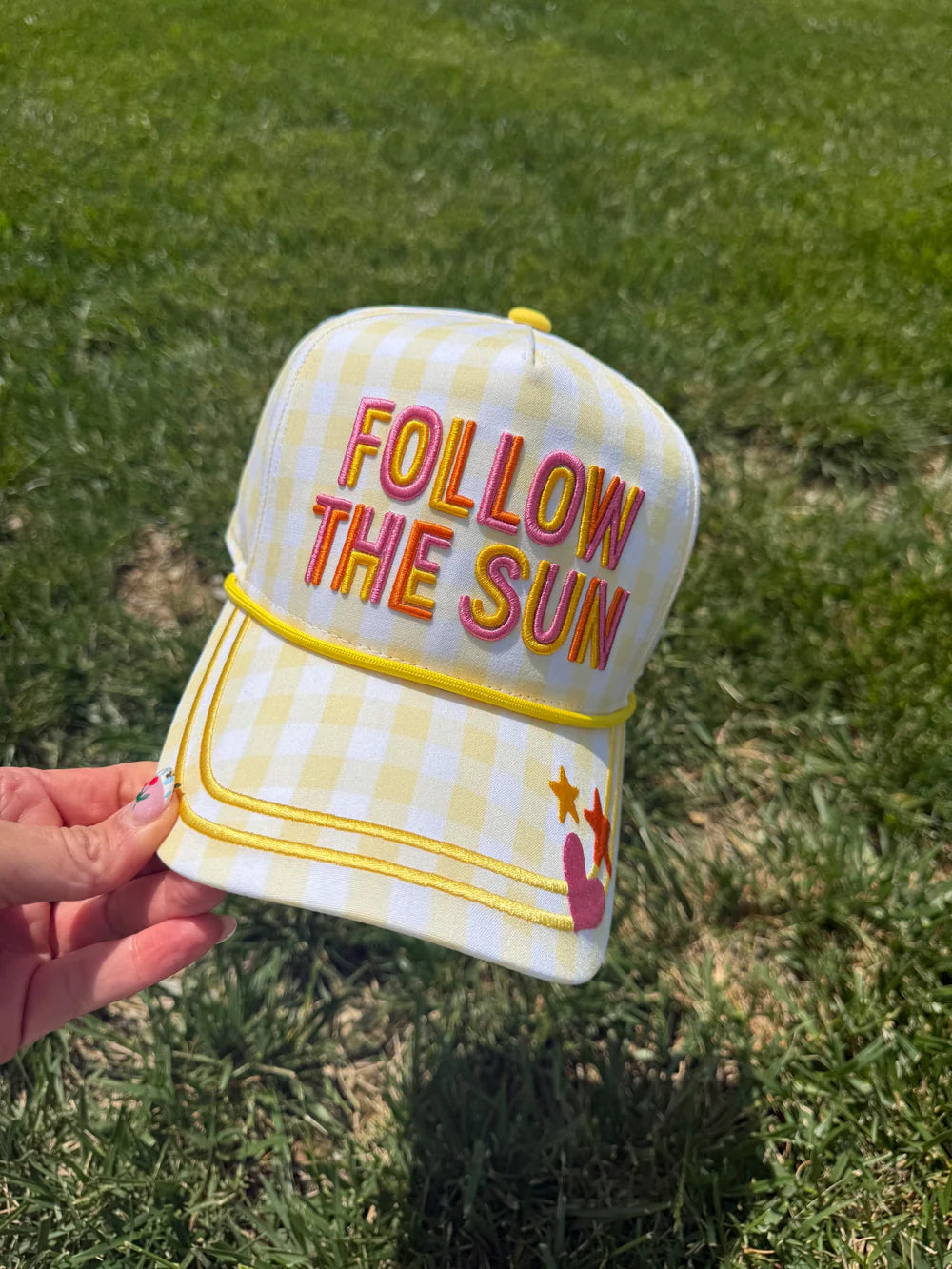 Hand holding hat embroidered with 'follow the sun' in the grass