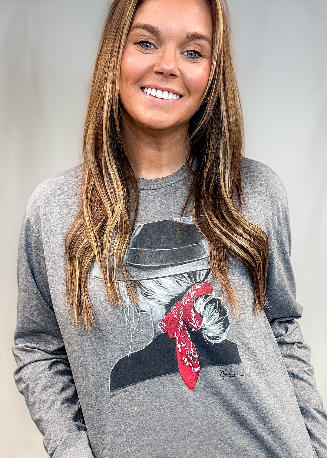 A smiling woman with long light brown hair wearing a heather gray long-sleeve graphic t-shirt with a cowboy hat and red bandana print, paired with blue jeans.
