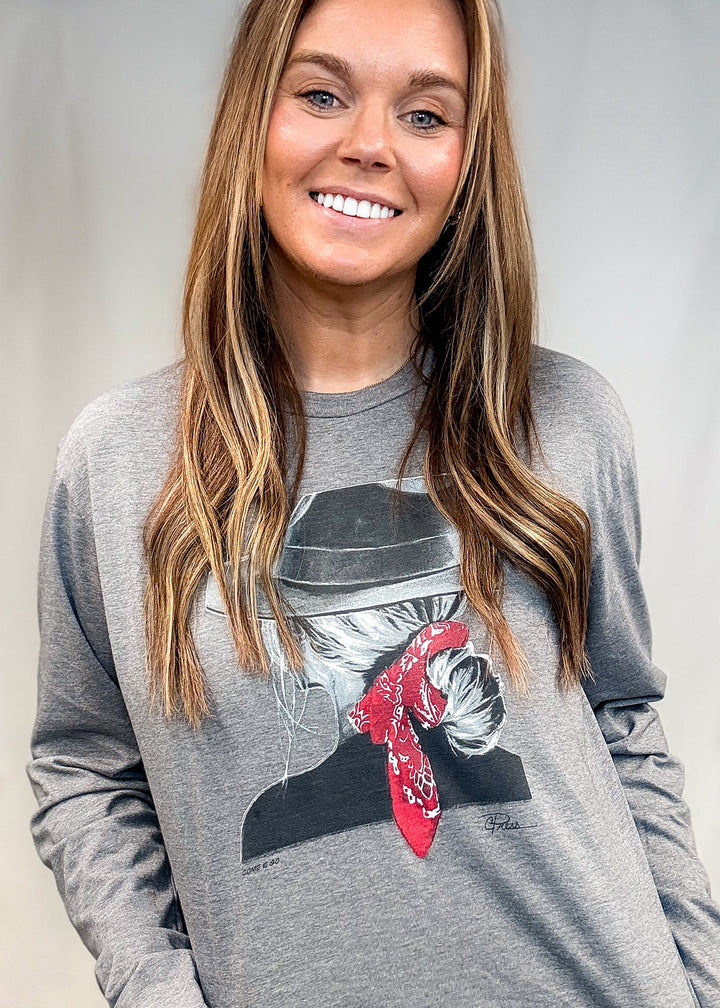 A smiling woman with long light brown hair wearing a heather gray long-sleeve graphic t-shirt with a cowboy hat and red bandana print, paired with blue jeans.