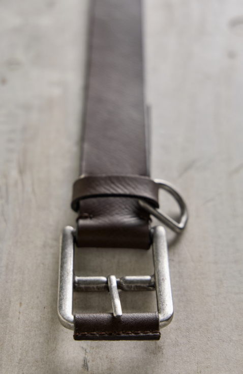 A close-up of a dark brown leather belt with a silver-toned metal buckle, laid on a textured light gray surface.