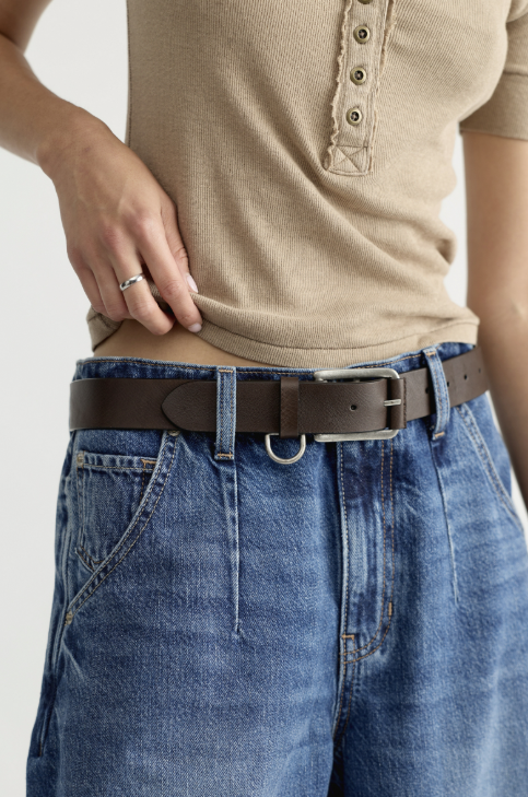 A close-up of a person wearing a dark brown leather belt with a silver buckle, paired with blue jeans and a beige short-sleeve henley top.