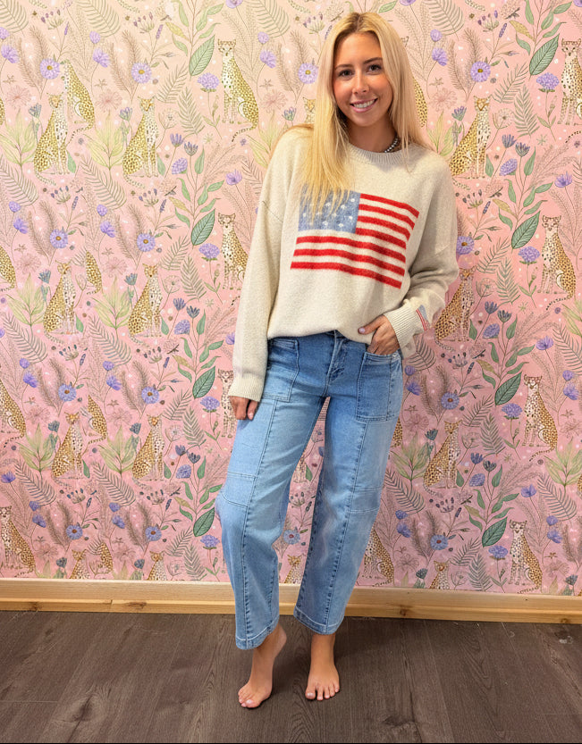 A woman wearing a cream knit sweater with a red, white, and blue American flag graphic on the chest, paired with light blue jeans, standing in a room with pink patterned wallpaper and wood flooring.