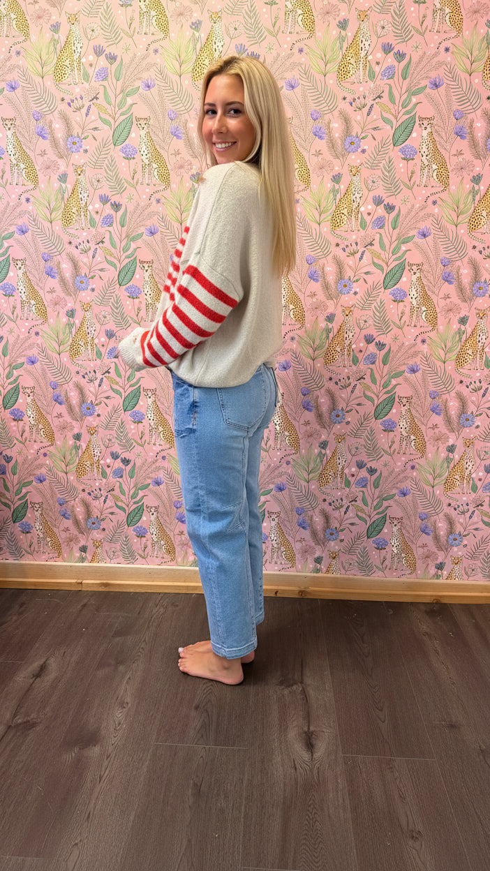A woman wearing a cream knit cardigan with red stripes on the sleeve, blue jeans, and bare feet, standing in a room with pink floral wallpaper and a dark wood floor.