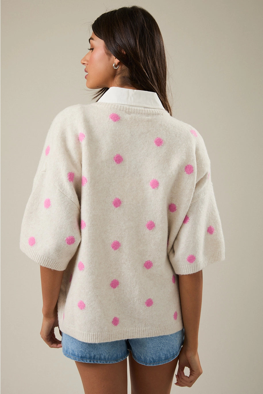 Woman wearing a beige cardigan with pink polka dots and denim shorts on a neutral background