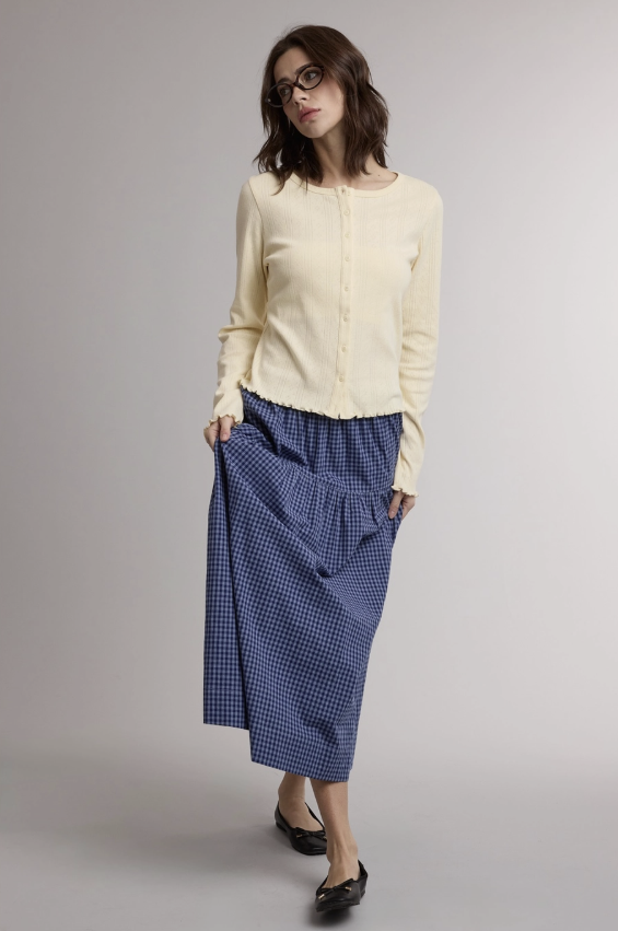 A woman modeling a cream long-sleeve button-front cardigan and a blue gingham midi skirt against a plain light gray studio background.