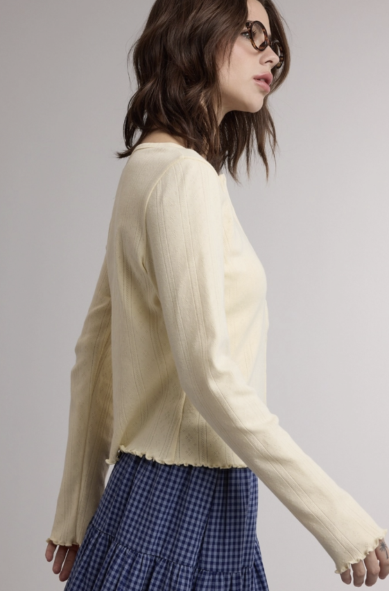 A woman wearing a cream ribbed knit long-sleeve cropped sweater and a blue-and-white gingham skirt, photographed against a plain light gray studio background.