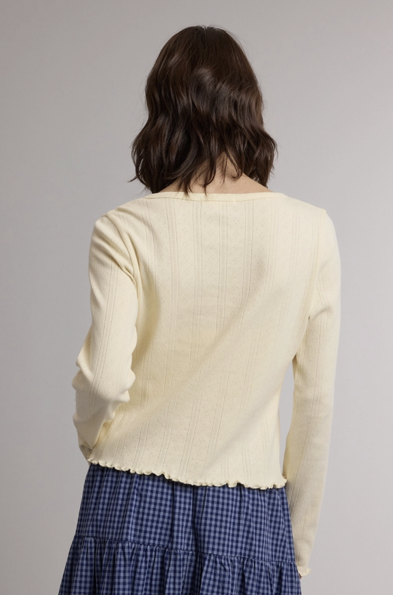 A woman shown from behind wearing a cream ribbed knit long-sleeve top with a scalloped hem and a blue gingham skirt, against a plain light gray studio background.
