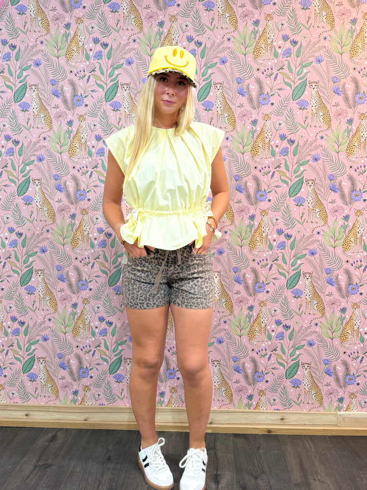 Person wearing a yellow top and patterned shorts standing against a pink floral and animal-patterned wall.