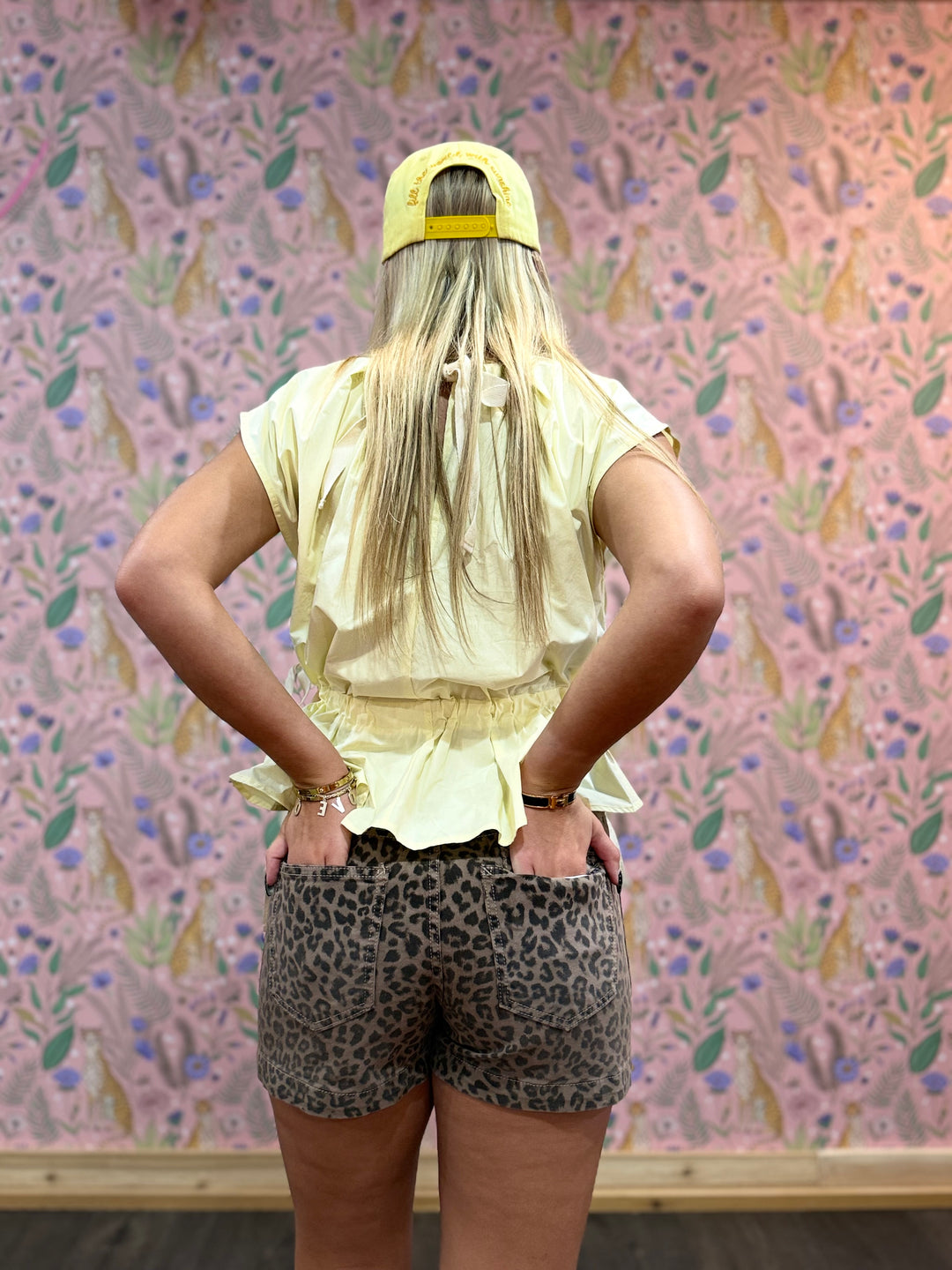 Person wearing a yellow cap and light-colored top with leopard print shorts in front of floral wallpaper.