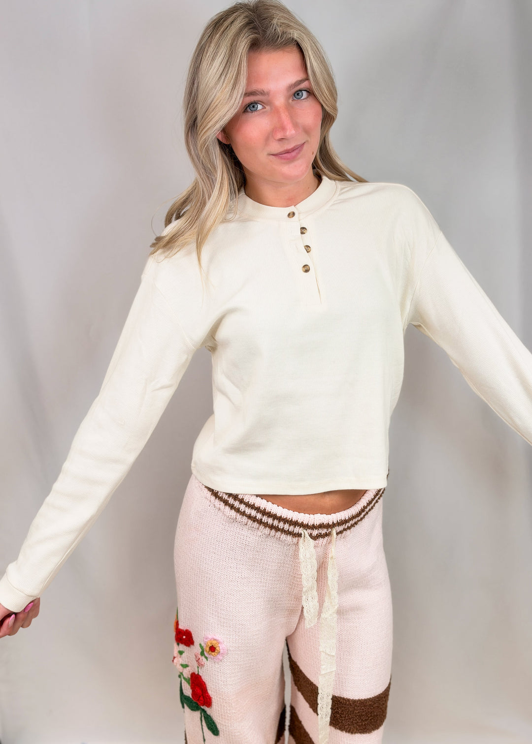 A woman modeling a cream long-sleeve henley crop top and light pink knit pants with brown stripes and red floral embroidery, posed against a light gray studio backdrop.
