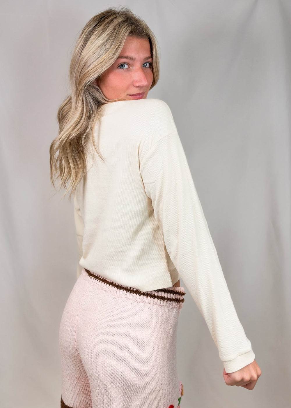 A woman with blonde hair is shown from the back wearing a cream-colored long-sleeve knit top and light pink knit pants with a brown patterned waistband, set against a light gray fabric backdrop.
