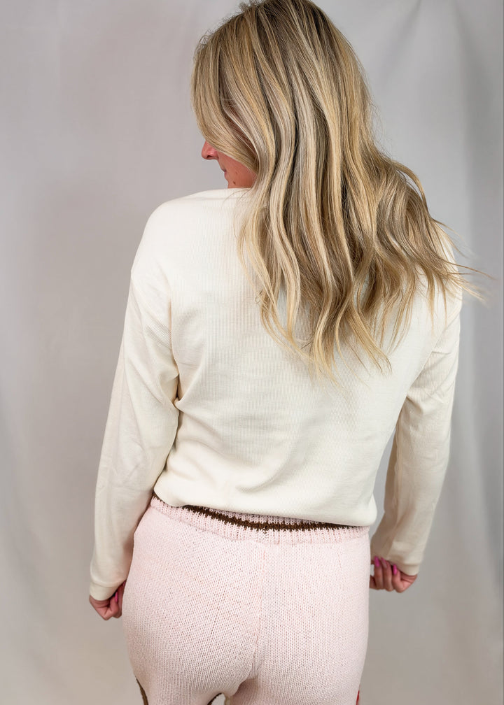 A woman shown from behind wearing a cream-colored long-sleeve knit top and light pink textured leggings against a neutral studio backdrop.