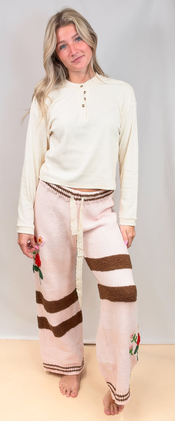 A woman stands barefoot against a plain light gray studio backdrop wearing a cream long-sleeve henley top and loose knit pajama pants in pale pink with brown stripes and small floral accents.
