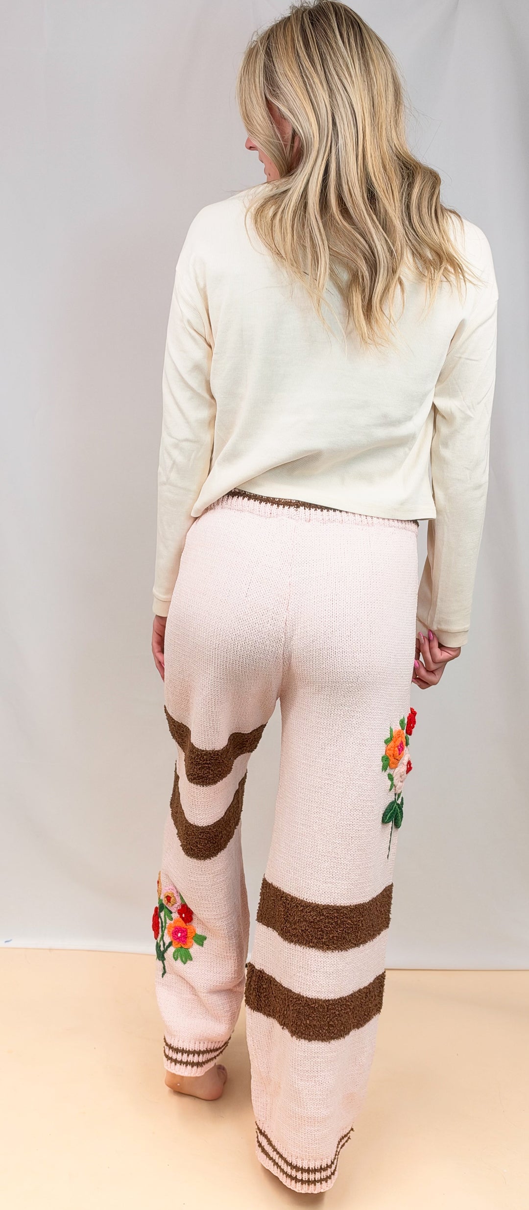 A woman viewed from behind wearing cream knit pants with brown horizontal stripes and colorful floral embroidery near the lower legs, paired with a cream long-sleeve top against a plain studio backdrop.